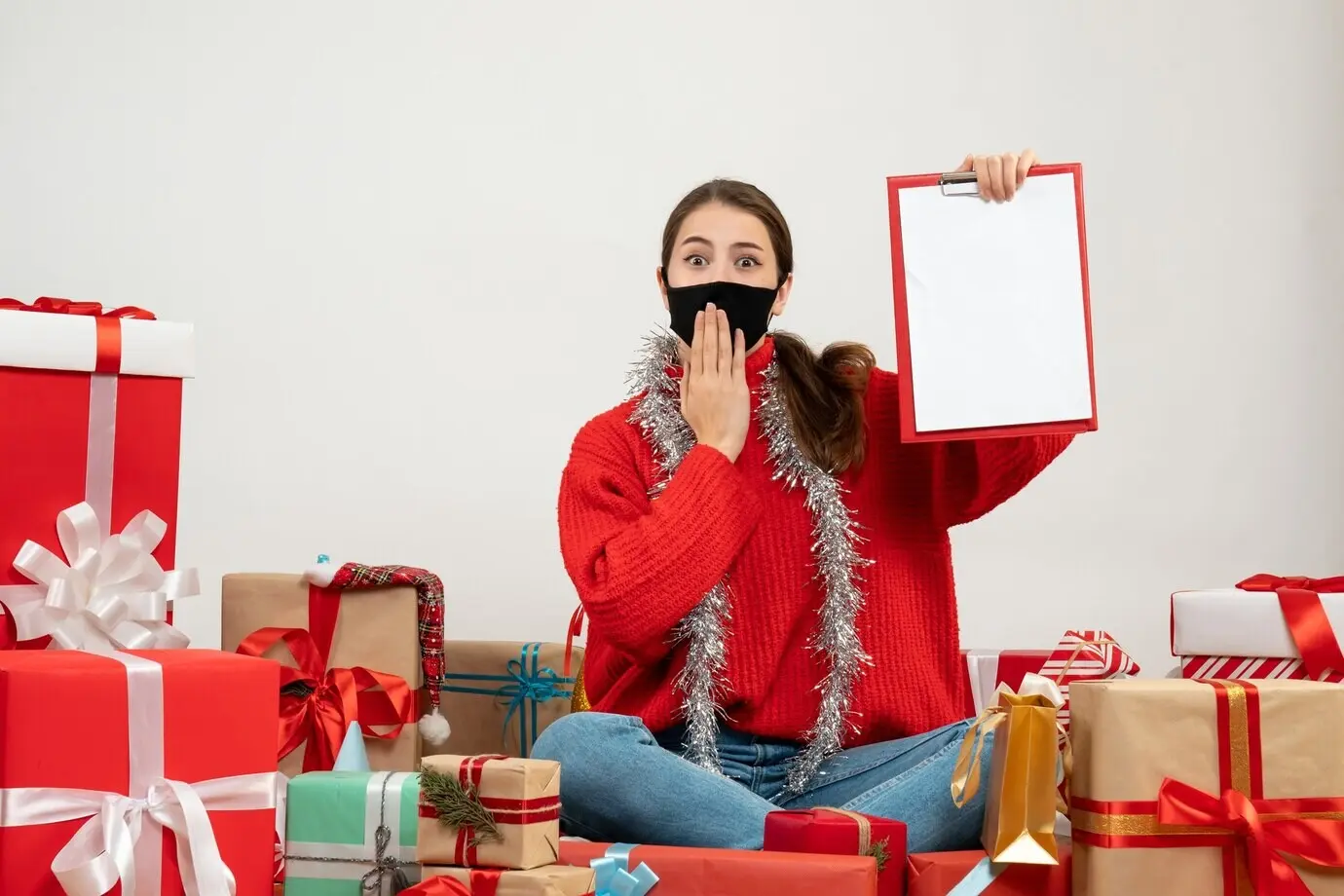 Удивленная девушка в черной маске, держащая документы, сидит среди подарков на белом фоне.